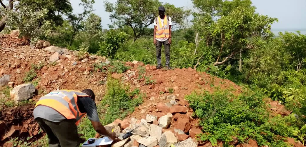 AMEA field team in high-visibility vests examining an exploration trench in red laterite soil